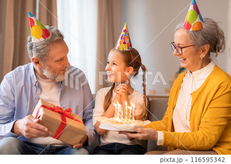 A young girl, adorned with a festive party hat, beams with joy as her grandparents, also wearing colorful party hats, present her with a birthday cake and a wrapped gift during an indoor celebration A young girl, adorned with a festive party hat, beams with joy as her grandparents, also wearing colorful party hats, present her with a birthday cake and a wrapped gift during an indoor celebration 116595342
