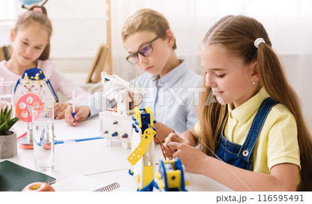 Three young students are sitting at a table in a classroom setting, working on building robots. They are concentrating on their work and seem to be enjoying the activity. 116595491