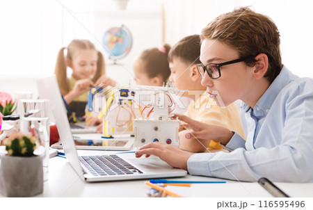 A teenage boy wearing glasses sits at a desk in a classroom, using a laptop computer. He is surrounded by other students, including a girl who appears to be working on a small robot. 116595496