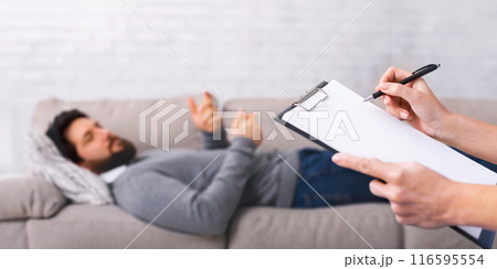 A young man lies comfortably on a gray couch, gesturing with his hands while speaking to someone off-camera. The listener holds a clipboard and pen, ready to take notes or provide feedback. A young man lies comfortably on a gray couch, gesturing with his hands while speaking to someone off-camera. The listener holds a clipboard and pen, ready to take notes or provide feedback. 116595554