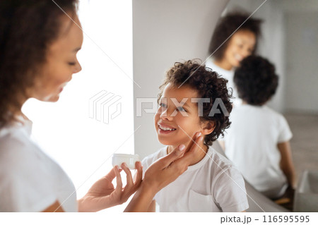 African American mother tenderly applies sunscreen to her child cheerful face, ensuring protection from the suns rays. Both are dressed in casual white attire, reflecting a carefree, sunny day. African American mother tenderly applies sunscreen to her child cheerful face, ensuring protection from the suns rays. Both are dressed in casual white attire, reflecting a carefree, sunny day. 116595595