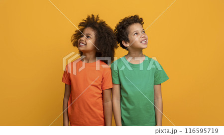 Two joyful African American children are standing side by side, one wearing an orange shirt and the other in green, both gazing upwards with happy expressions on their faces 116595719