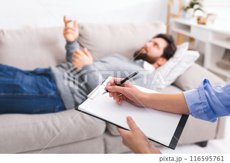 A man is lying on a couch, gesturing with his hand as he speaks, while engaged in a counseling session. A therapist, partially visible in the foreground, is attentively taking notes on a clipboard. 116595761