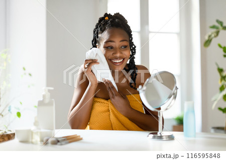 African American woman with dark braids wrapped in a yellow towel is wiping her face with a white washcloth while smiling at a mirror. She is sitting at a vanity table with makeup brushes 116595848