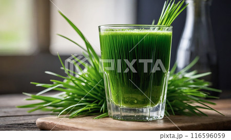 A glass of green wheatgrass juice with fresh blades of wheatgrass on a rustic wooden table, illuminated by soft light. 116596059