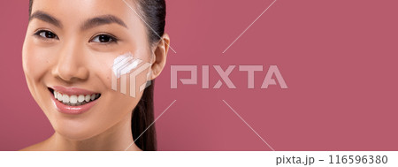 Closeup portrait of smiling beautiful young asian woman with moisturizer cream on her pretty face, testing new beauty product, purple studio background, copy space. Face care concept 116596380