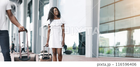 African American couple walks together outside a modern building with large windows, each pulling a suitcase. The couple is dressed casually in summer clothes, copy space African American couple walks together outside a modern building with large windows, each pulling a suitcase. The couple is dressed casually in summer clothes, copy space 116596406