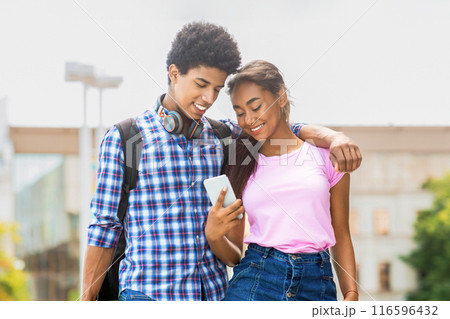 This image shows a teen black couple walking down a city street. Guy, wearing a plaid shirt and headphones, looking at his phone. His girlfriend, who is wearing a pink shirt and denim skirt, smiling 116596432