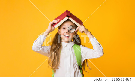 Cheerful Curious School Girl Looking Through Magnifier At Camera Covering Head With Book On Yellow Studio Background. Free Space Cheerful Curious School Girl Looking Through Magnifier At Camera Covering Head With Book On Yellow Studio Background. Free Space 116596464