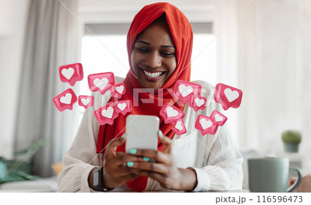 African American woman wearing a red hijab smiles while looking at her smartphone, surrounded by pink heart icons representing social media likes. She is sitting at a table in a bright, modern room. African American woman wearing a red hijab smiles while looking at her smartphone, surrounded by pink heart icons representing social media likes. She is sitting at a table in a bright, modern room. 116596473