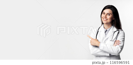 A smiling female doctor in a white coat points to the right with her right hand. Her arms are crossed and she is wearing a stethoscope. A smiling female doctor in a white coat points to the right with her right hand. Her arms are crossed and she is wearing a stethoscope. 116596591