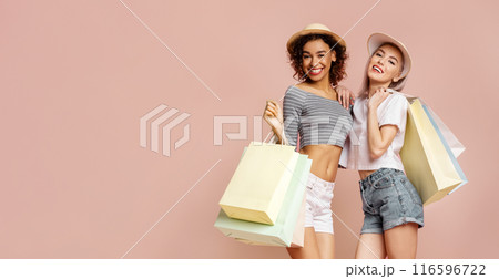 Two young women wearing summer outfits and hats are holding shopping bags while smiling at the camera. They are standing in front of a pink background, copy space Two young women wearing summer outfits and hats are holding shopping bags while smiling at the camera. They are standing in front of a pink background, copy space 116596722
