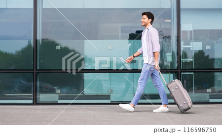 Vacations and travel concept. Young man walking with luggage suitcase at airport, side view with free space 116596750