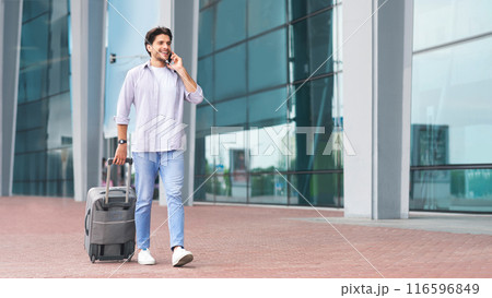 Calling taxi. Millennial man arriving at airport and ordering car by cell phone, walking with suicase outdoors near terminal, free space 116596849