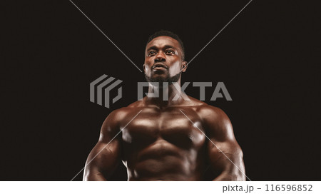 A muscular man stands against a black background, looking upwards with a determined expression. A muscular man stands against a black background, looking upwards with a determined expression. 116596852