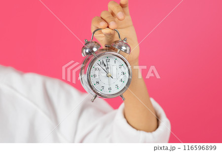 A hand is holding a silver alarm clock with the hands at 12 o'clock. The clock has a white face with black numbers and hands. The background is a bright pink, cropped A hand is holding a silver alarm clock with the hands at 12 o'clock. The clock has a white face with black numbers and hands. The background is a bright pink, cropped 116596899