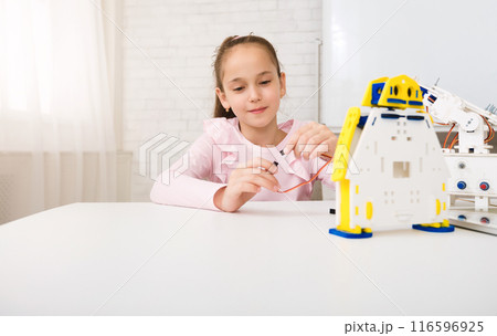 A young girl sits at a white table, focused on assembling a robot kit. She holds a small wire in her hand, carefully connecting it to the robots wiring. The robot is made of white plastic 116596925
