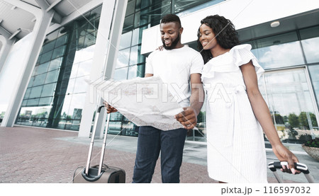 Black couple in casual white clothing stand outside an airport terminal, looking at a map, as they prepare to embark on their travels. 116597032