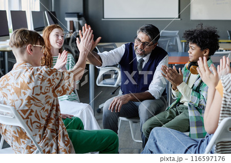 Cheerful mature school psychologist doing high five with male student during group therapy session, other teenagers clapping hands Cheerful mature school psychologist doing high five with male student during group therapy session, other teenagers clapping hands 116597176