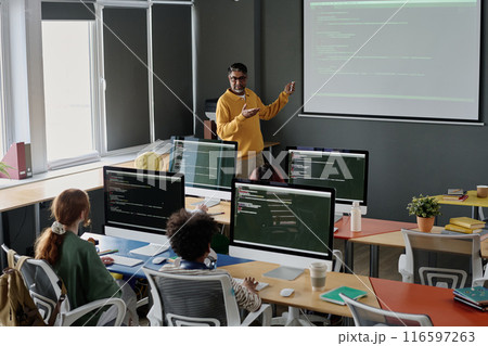 Mature biracial teacher of Computer Science explaining subject to group of teenagers during lesson at school 116597263