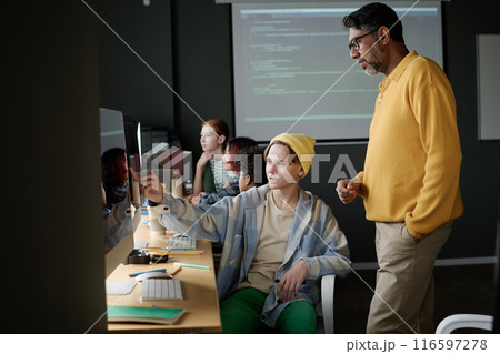 Caucasian teenager wearing casual clothes sitting at desk showing code on computer screen to his teacher, medium long shot 116597278