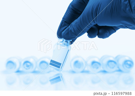 Blue gloved doctor hand holding a vaccine vial in front of a blurred row of vaccine vials on a blue background. Blue gloved doctor hand holding a vaccine vial in front of a blurred row of vaccine vials on a blue background. 116597988