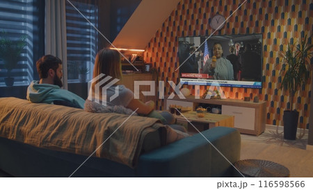 Couple watches news about United States elections on TV screen sitting on sofa Couple watches news about United States elections on TV screen sitting on sofa 116598566