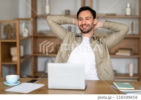 A young man with a cheerful expression sits back at his desk in a well-lit, contemporary office setting, hands clasped behind his head. In front of him is an open laptop, alongside a cup of coffee 116599658
