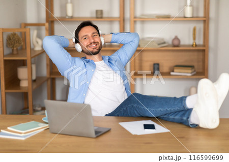 A man is seated at a desk, wearing headphones and working on a laptop placed on his lap. He appears focused and engaged in his work, with various papers and stationery scattered around the desk. A man is seated at a desk, wearing headphones and working on a laptop placed on his lap. He appears focused and engaged in his work, with various papers and stationery scattered around the desk. 116599699