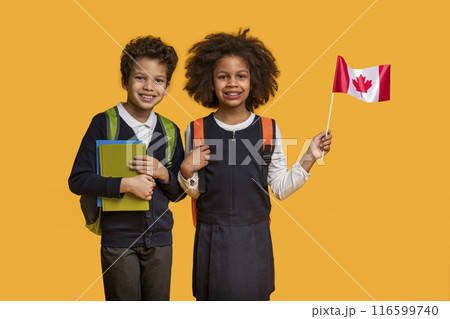 African American boy and girl, both wearing school backpacks and casual clothing, stand side by side with bright smiles. The girl is holding a small Canadian flag, and both are equipped with notebooks African American boy and girl, both wearing school backpacks and casual clothing, stand side by side with bright smiles. The girl is holding a small Canadian flag, and both are equipped with notebooks 116599740