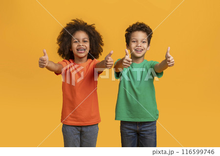 Two African American young kids, one boy and one girl, are enthusiastically giving thumbs up gestures while smiling against a vibrant orange background 116599746