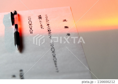白いテーブルに置かれた手書きの退職届とゲルインクの黒ペンを若干赤みがかった照明をかけて撮ったシーン 116600212