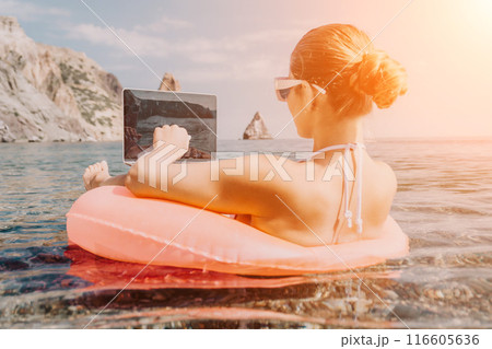 Woman freelancer works on laptop swimming in sea on pink inflatable ring. Happy tourist in sunglasses floating on inflatable donut and working on laptop computer in calm ocean. Remote working anywhere 116605636