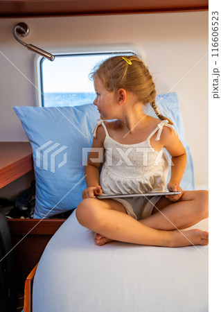 Little girl sits with tablet in the cabin of a yacht and looks out the window during a sea crossing. Yacht life concept 116606523