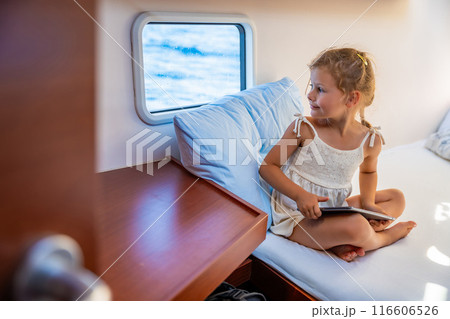 Little girl sits with tablet in the cabin of a yacht and looks out the window during a sea crossing. Yacht life concept Little girl sits with tablet in the cabin of a yacht and looks out the window during a sea crossing. Yacht life concept 116606526
