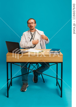 Man, doctor in white coat sitting at desk with laptop and books, using stethoscope, leading online consultation against cyan background Man, doctor in white coat sitting at desk with laptop and books, using stethoscope, leading online consultation against cyan background 116607640