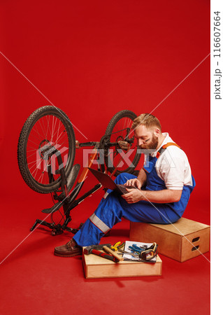 Bearded young man in blue overalls, mechanic sitting next to bicycle and looking on laptop against red background 116607664