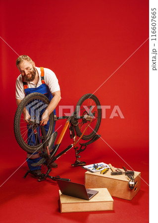Mechanic in blue overalls, bearded man working on bicycle, surrounded by various tools and laptop against red background 116607665