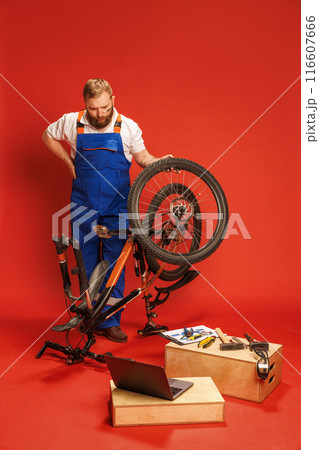Serious bearded young man in blue overalls repairing bicycle in workshop setting, with tools and laptop on wooden boxes against red background Serious bearded young man in blue overalls repairing bicycle in workshop setting, with tools and laptop on wooden boxes against red background 116607666