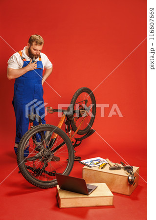 Bearded man, mechanic in blue overalls, with dirty face standing and looking on bike with thoughtful face, with laptop and tools around, against red background Bearded man, mechanic in blue overalls, with dirty face standing and looking on bike with thoughtful face, with laptop and tools around, against red background 116607669