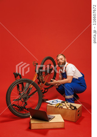 Mechanic, bearded man in blue overalls fixing bicycle, surrounded by tools and laptop, against red background 116607672