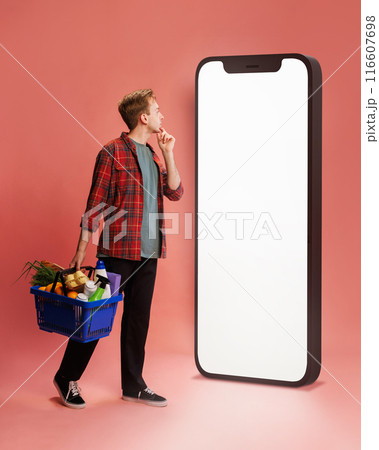 Thoughtful young man in checkered shirt, with basket of groceries looking at empty smartphone screen against pink background. Store website 116607698