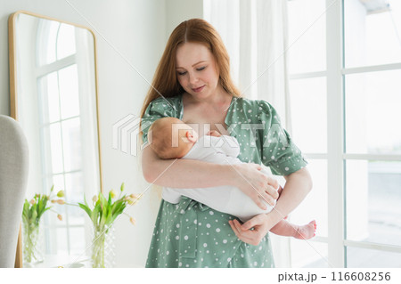Happy family at home. Mother breastfeeding baby for health nutrition wellness. Mom nursing feeding her newborn child milk at home. Woman infant girl in white bedroom Mother hugs baby with love care 116608256