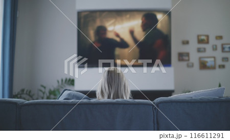 Woman sits on sofa and watches action movie on TV Woman sits on sofa and watches action movie on TV 116611291