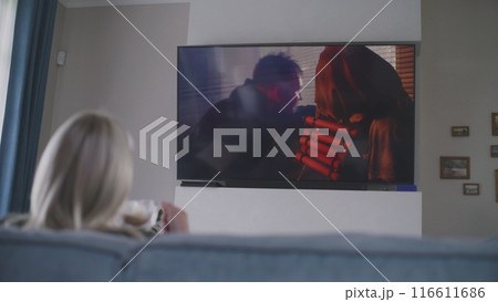 Woman sits on sofa and watches action movie on TV 116611686