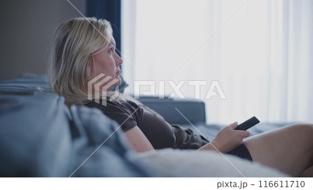 Side view of woman watching TV sitting on sofa and uses remote control Side view of woman watching TV sitting on sofa and uses remote control 116611710