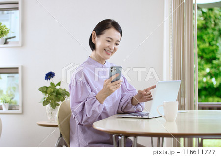 リビングでスマホを操作しながらパソコンを見るミドル女性 116611727