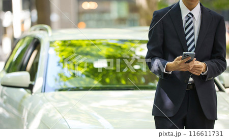 スマホを手に車の前に立つスーツの若い男性 スマホを手に車の前に立つスーツの若い男性 116611731