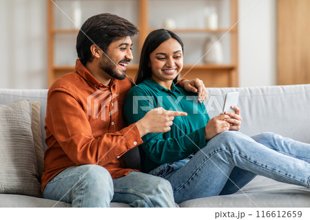 Indian man and woman are seated on a modern couch, engrossed in looking at a cell phone screen. They appear focused and engaged with the device, possibly browsing or communicating. Indian man and woman are seated on a modern couch, engrossed in looking at a cell phone screen. They appear focused and engaged with the device, possibly browsing or communicating. 116612659