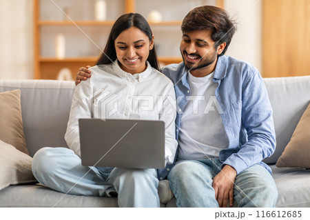 Indian man and woman are seated on a couch, both actively engaged in viewing content on a laptop screen. They appear focused and attentive, perhaps working or watching something together. 116612685